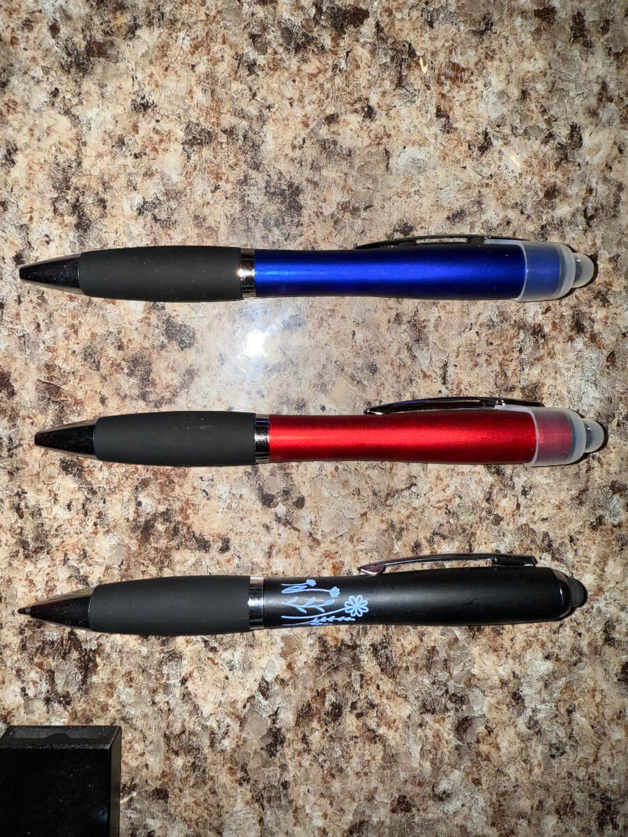 Set of three multi-functional stylus pens in blue, red, and black with black grips, LED light tips, and custom engraving, displayed on a countertop – ideal for business promotions, gifts, and personal use
