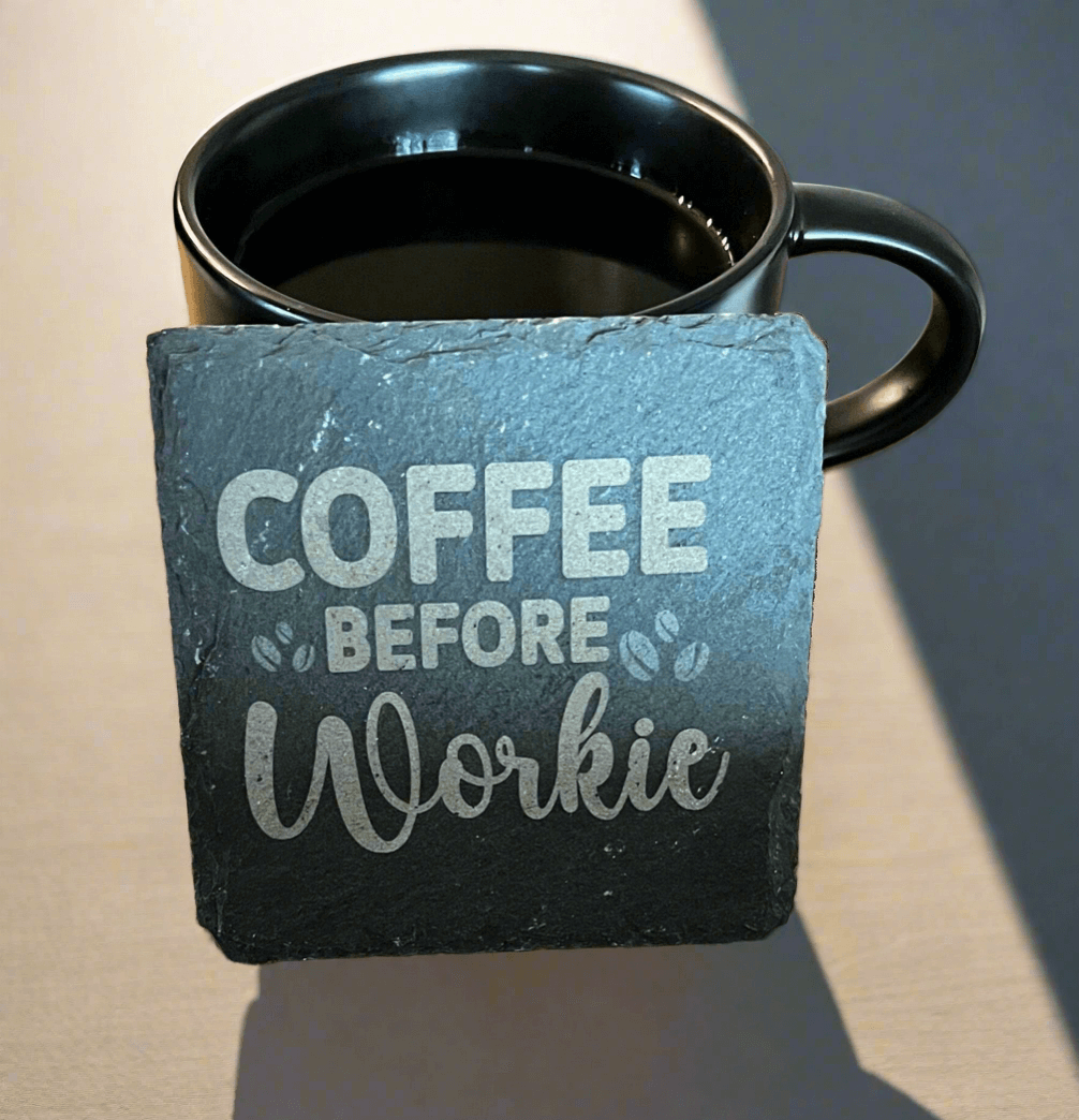 Black slate coaster engraved with the phrase “Coffee Before Workie” in bold and script fonts, placed in front of a black coffee mug on a light-colored table, personalized stone drinkware accessory for home or office.