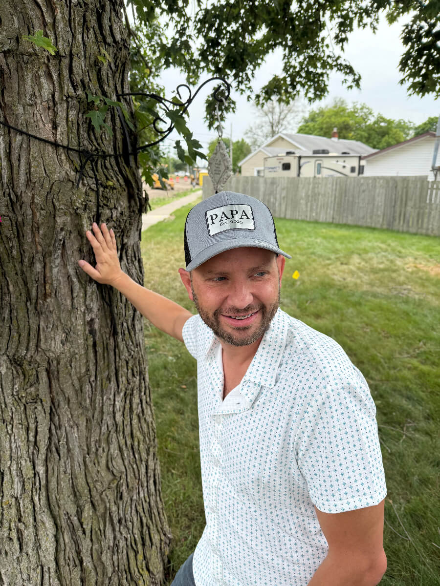 Man wearing a gray and black trucker hat with a personalized engraved leather patch that reads “PAPA Est. 2025,” standing outdoors by a tree and smiling, casual gift for new dads or grandfathers.