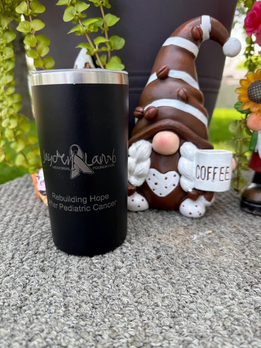 Engraved black 12oz tumbler with Jayden Lamb Memorial Foundation logo and rebuilding hope after pediatric cancer message next to coffee gnome decor
