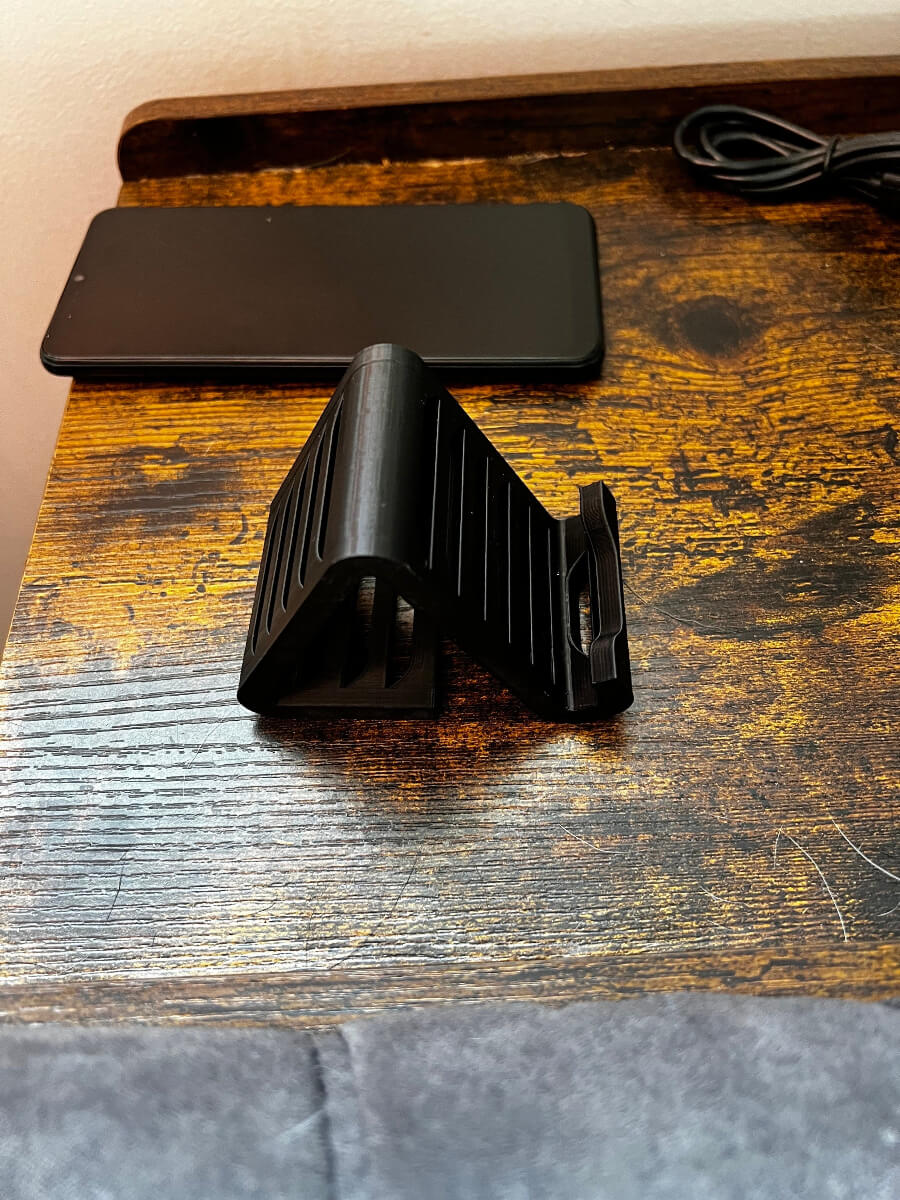 Black 3D printed phone stand made from PLA with a sleek, angled design, placed on a rustic wooden desk next to a smartphone and charging cable, custom desktop accessory for holding mobile devices.