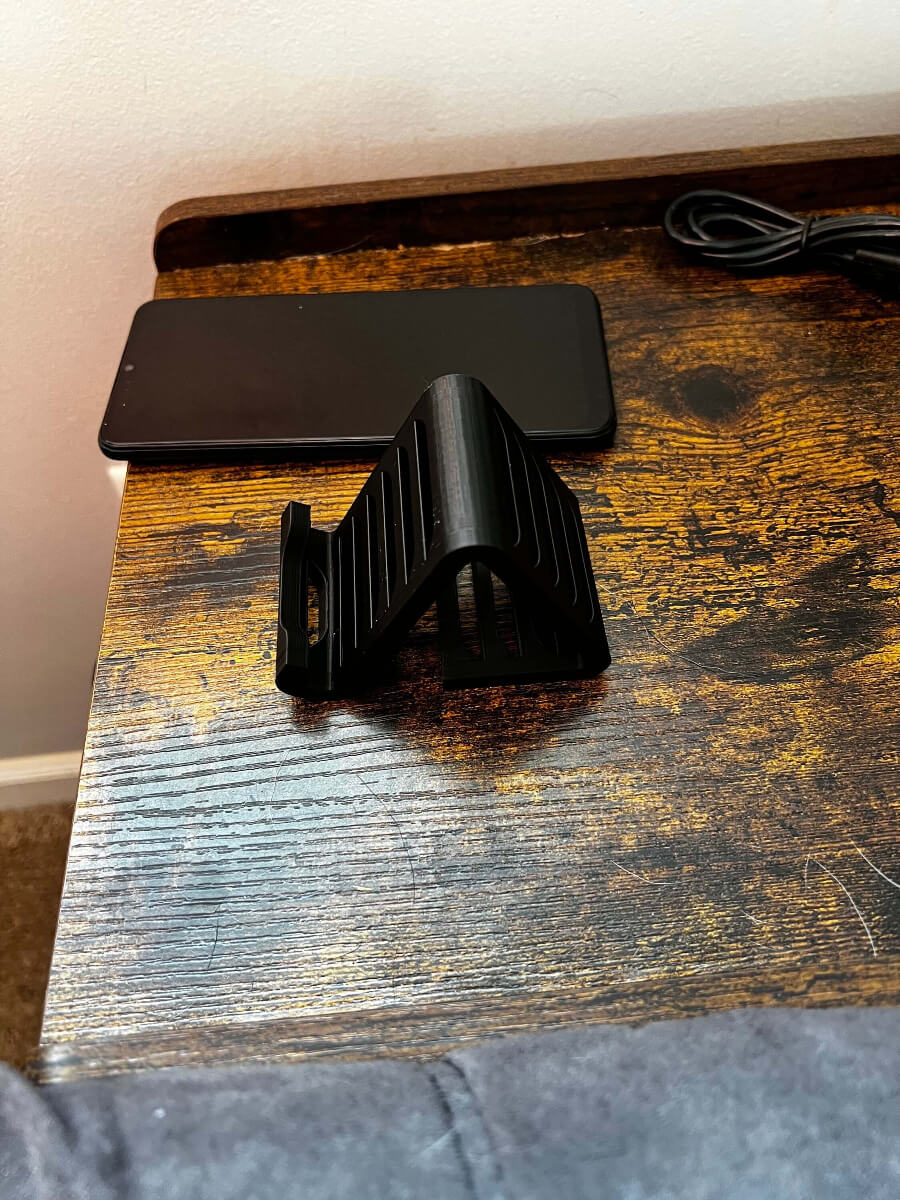Black 3D printed personalized phone stand with a modern, angled design placed on a rustic wooden desk next to a smartphone and a charging cable, custom desk accessory for holding mobile devices.