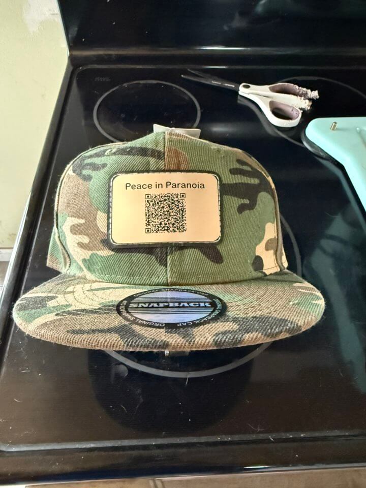 Camouflage snapback hat featuring a rectangular patch with the text “Peace in Paranoia” and a QR code, displayed on a stovetop