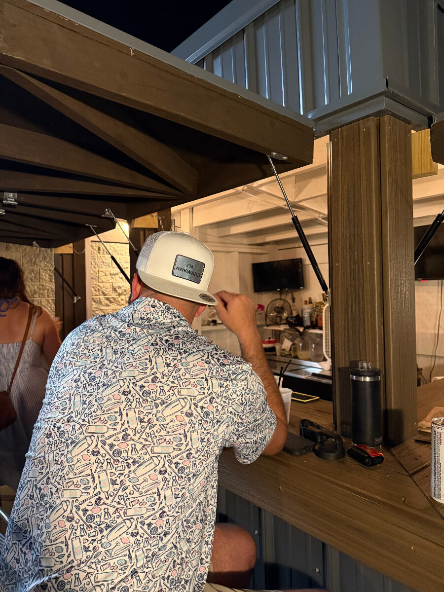 Person wearing a white trucker hat with “I’m Awkward” patch, seated at a counter in a casual indoor setting