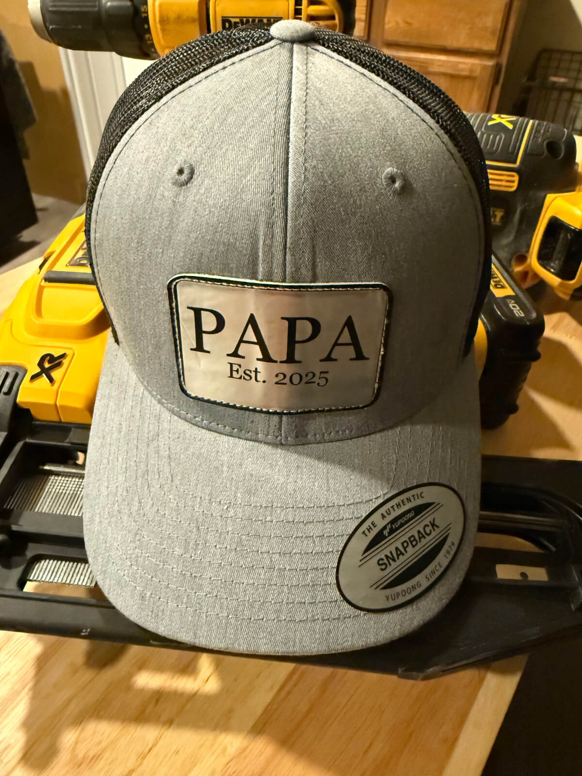Gray snapback trucker hat with a personalized engraved leather patch reading “PAPA Est. 2025,” displayed on a workbench with yellow power tools in the background, custom gift for new dads or grandfathers.
